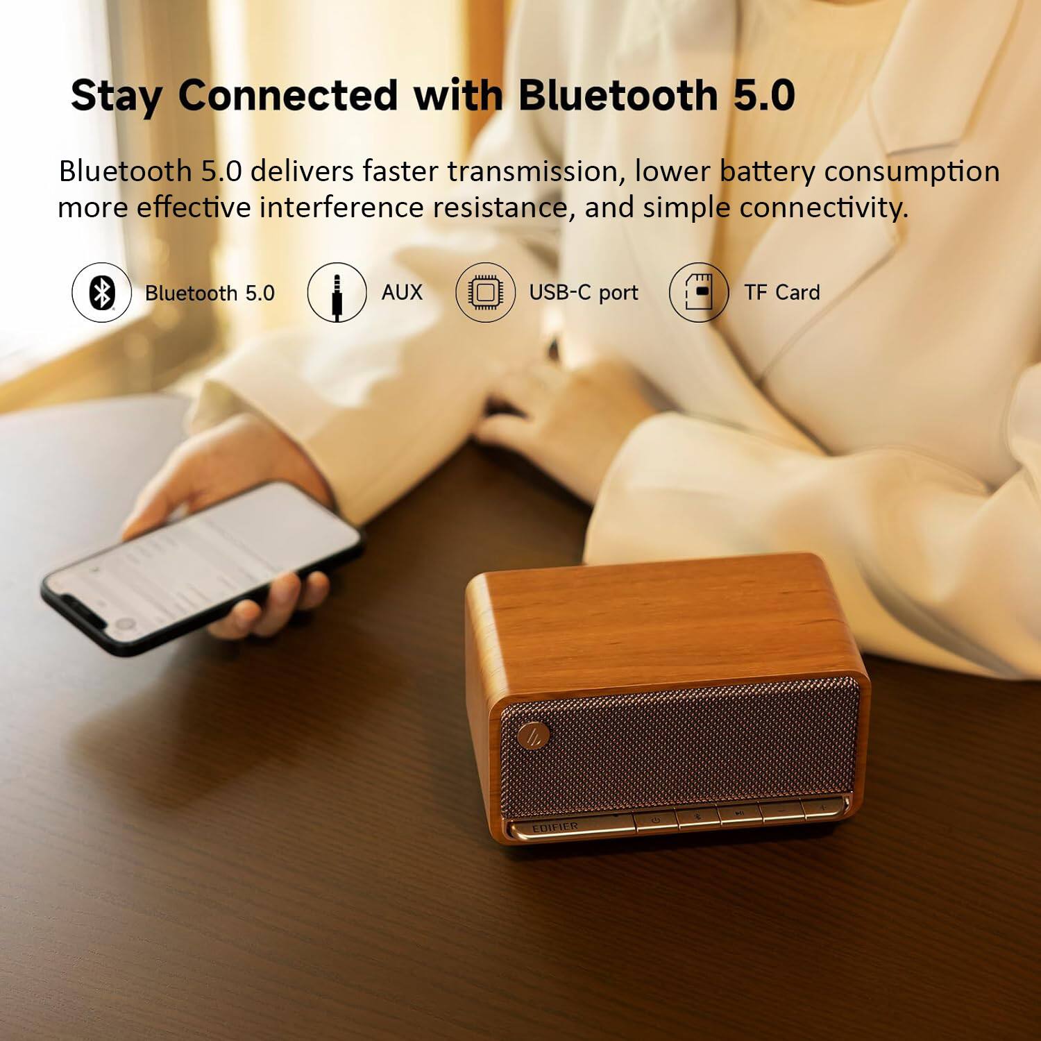 The image features a person holding a smartphone and a Bluetooth speaker on a table. The speaker is connected to the smartphone, and the person is likely using the speaker for better sound quality or to share the audio with others. The Bluetooth speaker is equipped with Bluetooth 5.0 technology, which offers faster transmission, lower battery consumption, more effective interference resistance, and simple connectivity. The speaker also has an AUX, USB-C port, and TF Card, providing multiple options for connecting and playing audio.