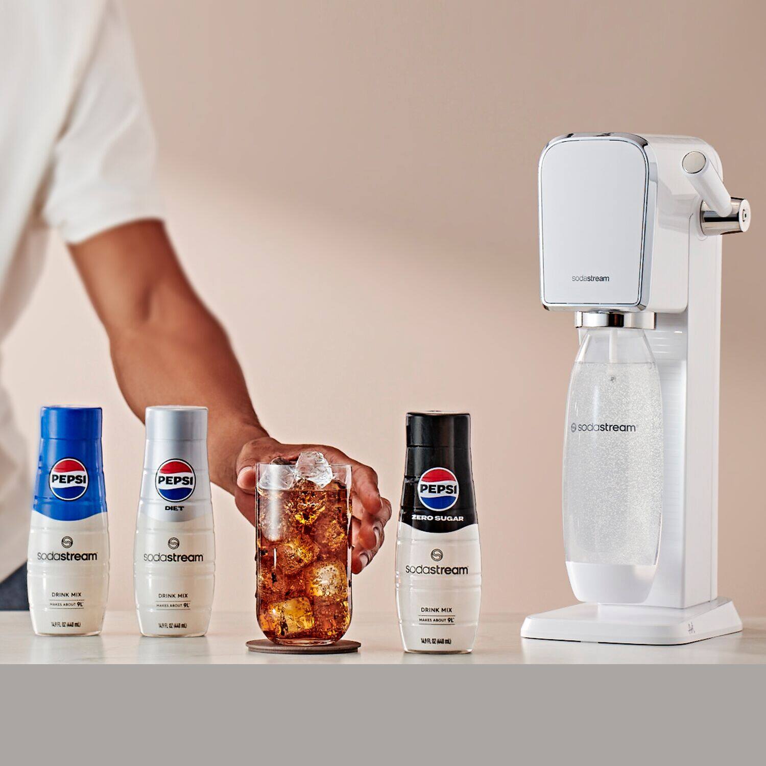 The image features a person holding a glass filled with ice and a carbonated drink, likely Pepsi, in front of a SodaStream machine. The machine is placed on a table, along with several bottles of Pepsi, which are lined up next to the machine. The scene suggests that the person is preparing to make a carbonated drink using the SodaStream machine.