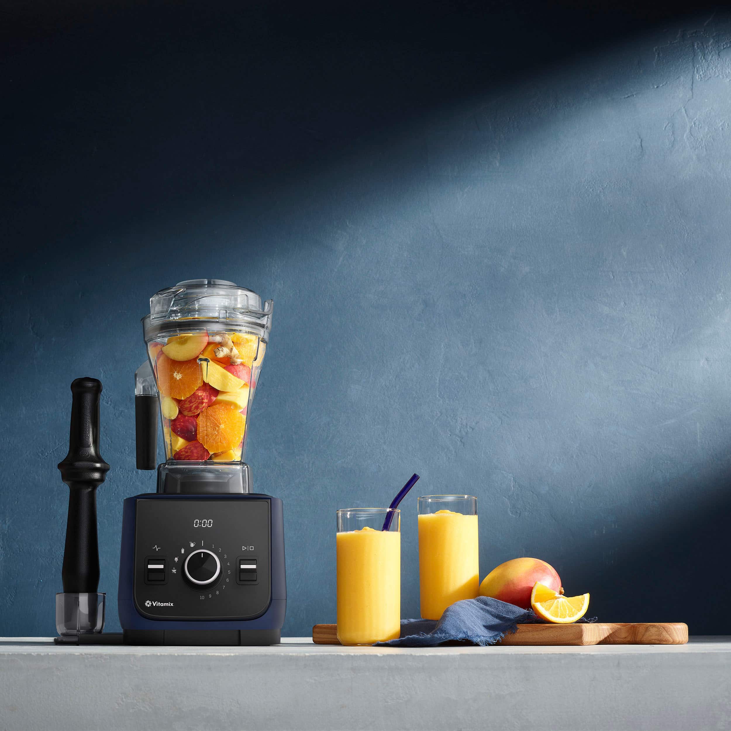 The image features a blue blender filled with fruit, including apples and oranges. There are two glasses of orange juice next to the blender, and a lemon is also present on the counter. The blender is placed on a countertop, and the scene is set against a dark blue wall.