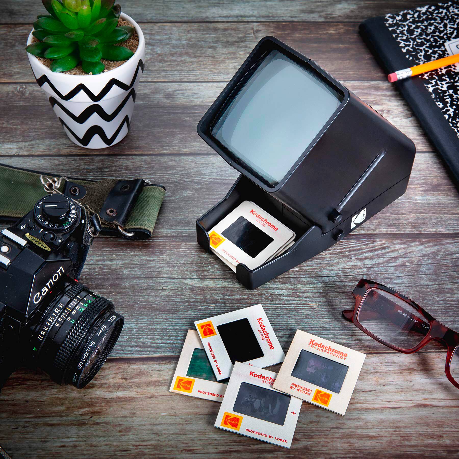 The image features a table with a variety of items, including a camera, a Kodachrome slide, a Kodachrome slide processor, and a potted plant. There are also some Polaroid pictures and a pair of glasses on the table. The table appears to be made of wood, and the items are arranged in a way that showcases the different types of photography equipment and accessories.