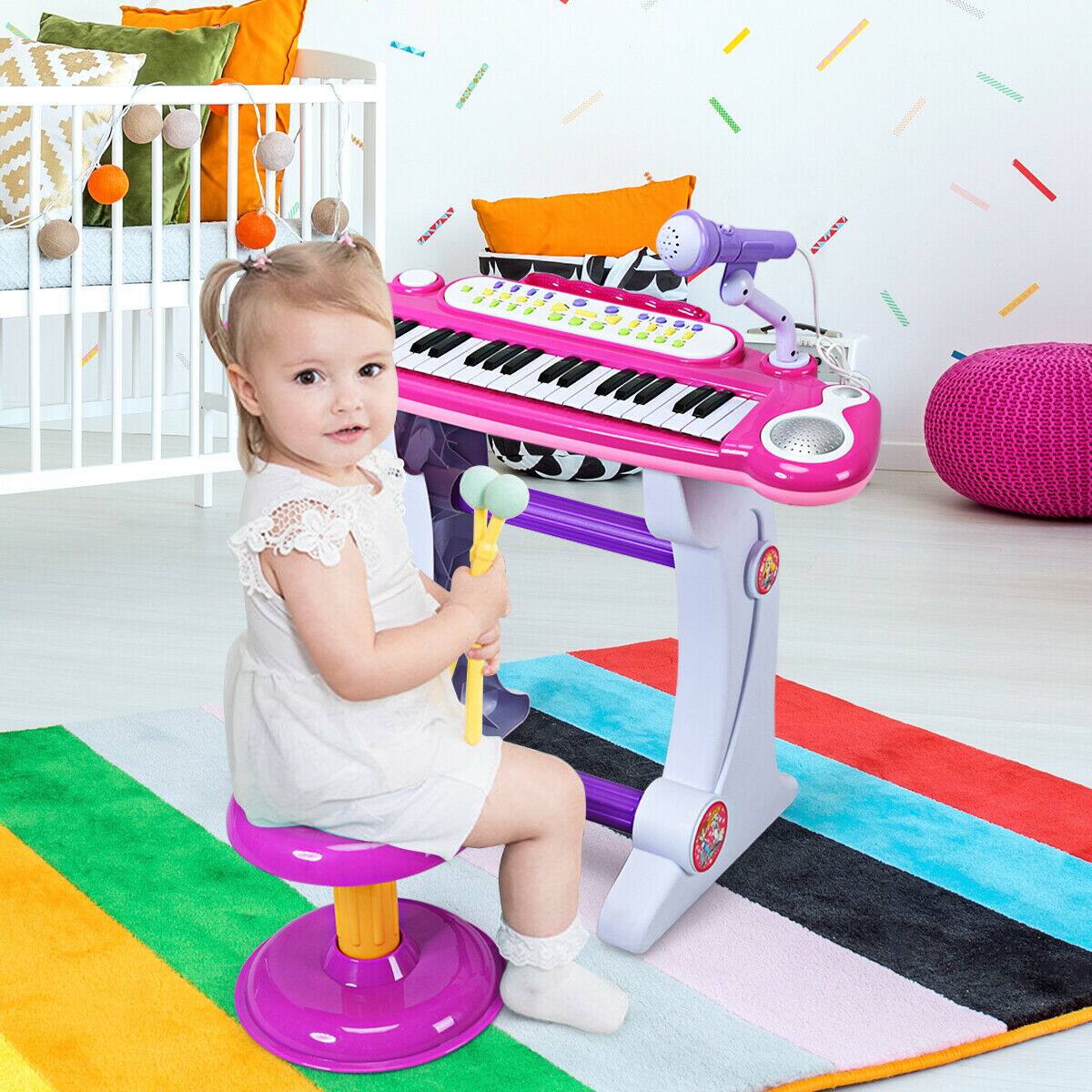 The image shows a little girl sitting on a purple stool in front of a pink keyboard. She is playing with the keyboard, which is placed on a colorful carpet. The scene is set in a room with a bed and a couch in the background. The girl appears to be enjoying her time playing with the keyboard.