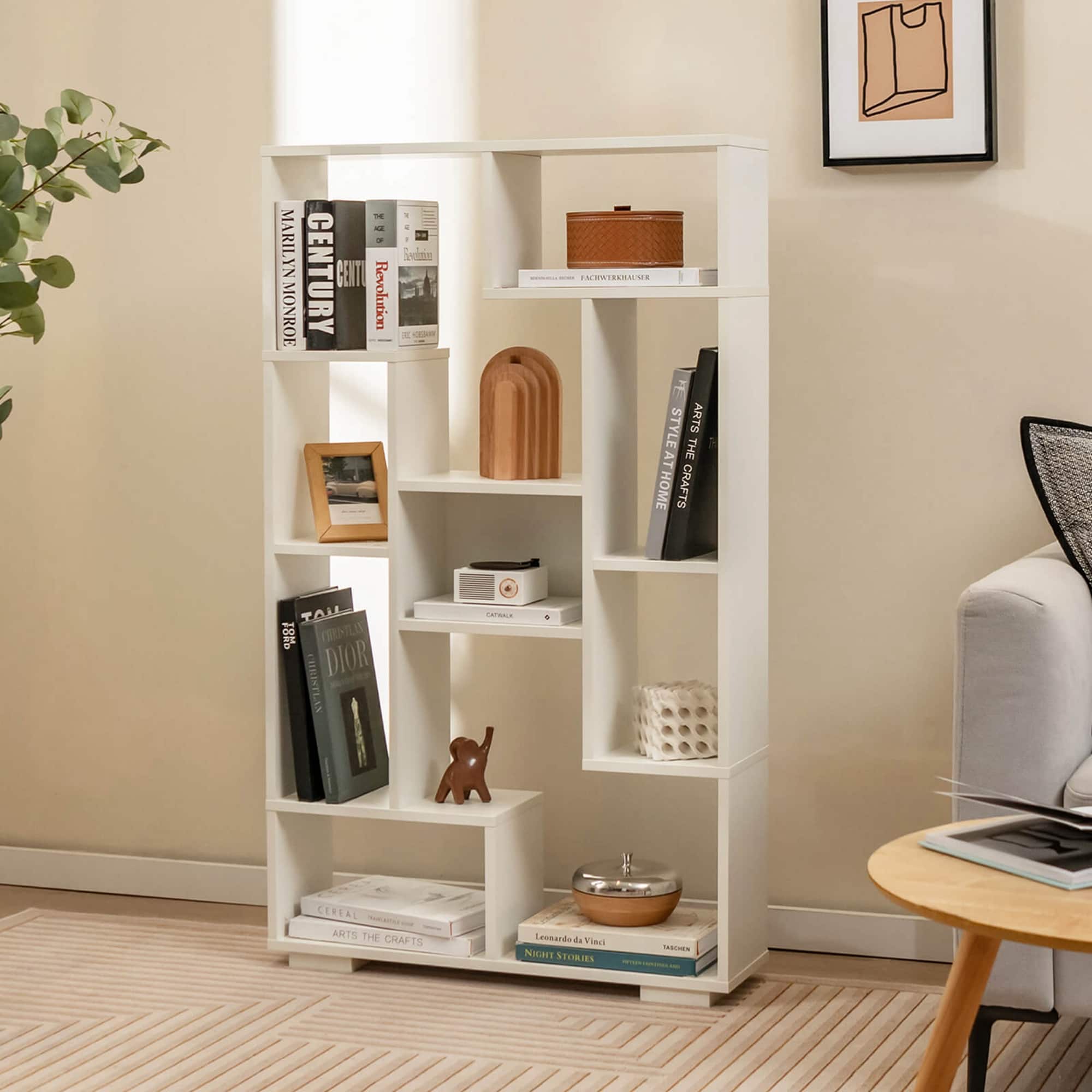 The image features a white bookshelf filled with various books and items. On the shelves, there are several books, including a book titled "Marilyn Monroe." Additionally, there are two bowls placed on the shelf, one near the center and the other towards the right side. A vase can also be seen on the shelf, located towards the left side. The bookshelf is situated in a room with a couch and a potted plant nearby.