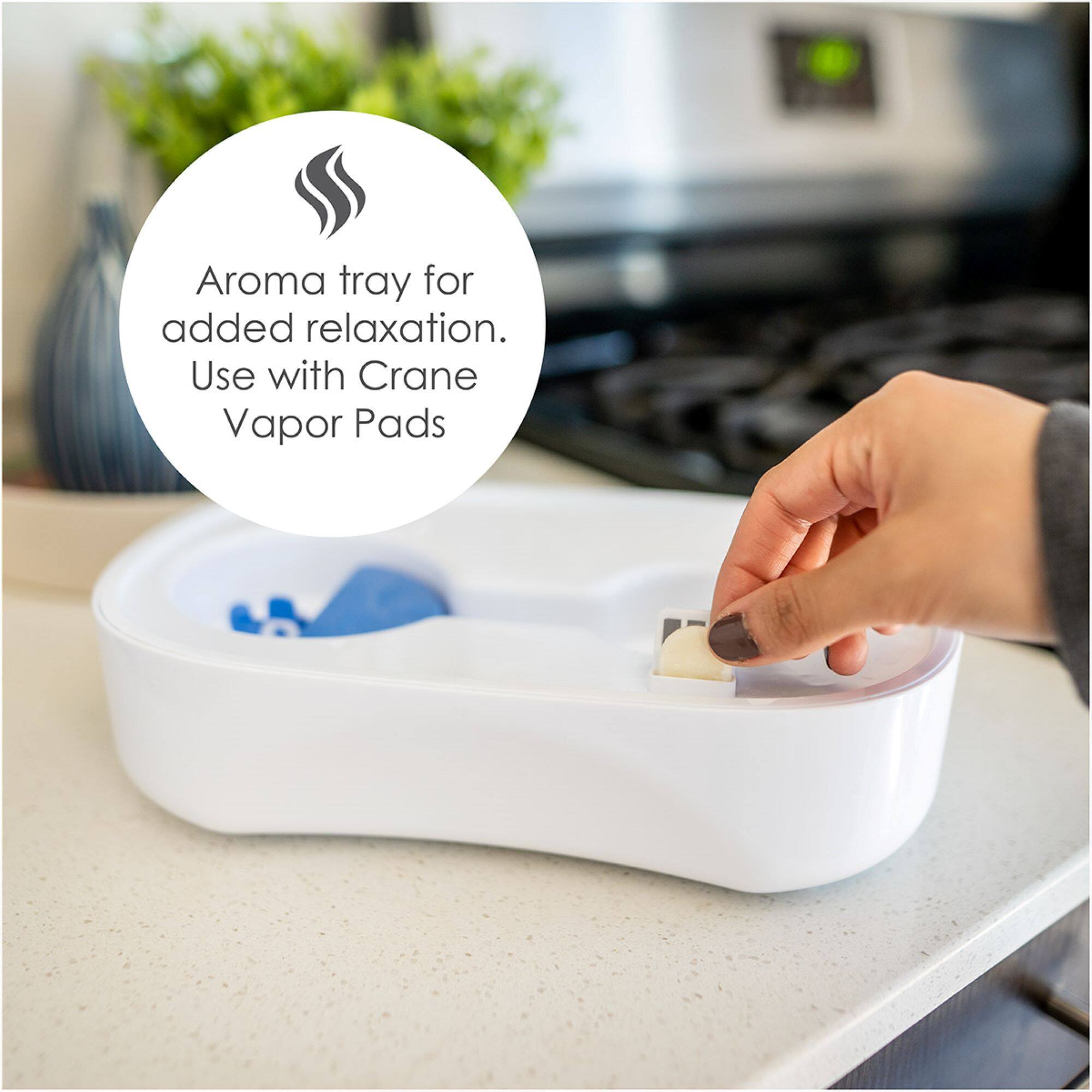 The image shows a person using an aroma tray for added relaxation, along with Crane Vapor Pads. The person is placing a pad into the tray, which is placed on a countertop. The aroma tray is designed to enhance relaxation and provide a pleasant fragrance experience.