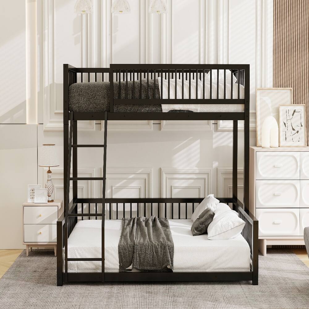The image shows a modern, black metal bunk bed in a well-lit, elegant room. The bed features a ladder on the left side, with the top bunk having a gray blanket and pillows, and the bottom bunk having white bedding with a gray throw blanket and pillows. The room has white paneled walls, a white dresser with drawers, and a small bedside table with a lamp and a book. There are also framed artworks on the dresser.