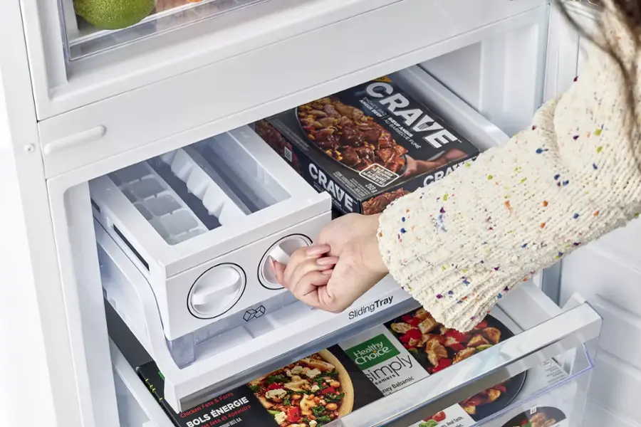 The image shows a person opening a refrigerator and pulling out a tray of food. The tray contains several items, including a pizza, a bowl of food, and a book. The person is reaching for the tray, which is placed on the bottom shelf of the refrigerator. The refrigerator is filled with various food items, making it a well-stocked kitchen.