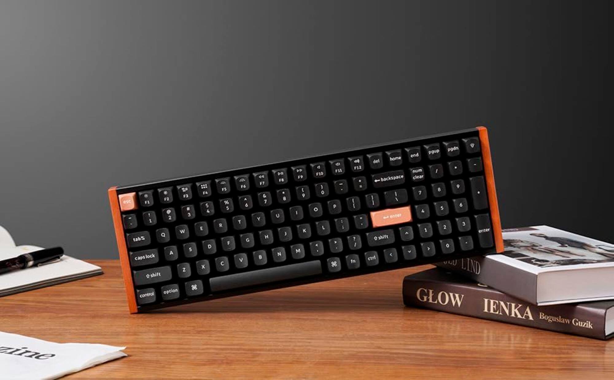 The image features a wooden desk with a keyboard and a book. The keyboard is placed on top of the book, which is titled "Glow". The keyboard is an orange and black computer keyboard, and it is positioned in such a way that it appears to be standing up. The book is placed on the left side of the keyboard, and the desk occupies the majority of the image.