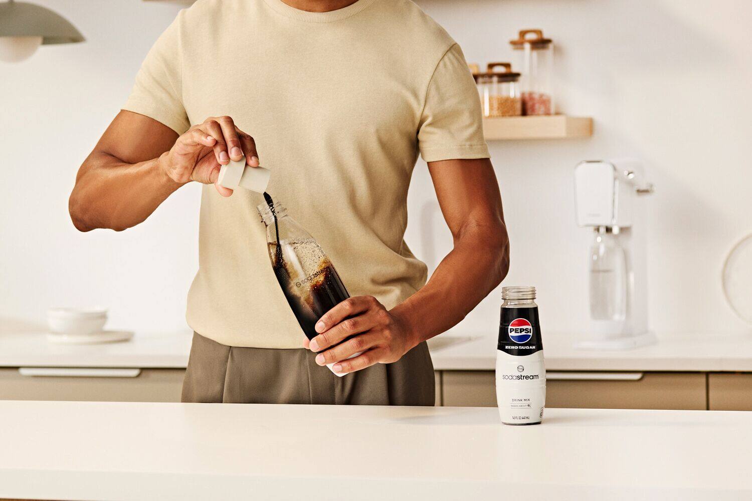 The image shows a man pouring a dark liquid, likely soda, into a glass bottle. The bottle is placed on a countertop, and there is a Pepsi logo visible in the scene. The man is using a soda stream to fill the bottle, and there are several other bottles nearby, possibly containing different types of soda.
