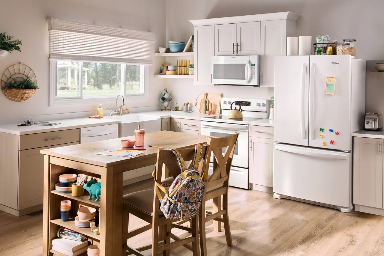The image shows a large kitchen with a wooden dining table surrounded by chairs. The kitchen is equipped with a refrigerator, microwave, oven, and sink. There are also various items such as bowls, bottles, and books scattered throughout the space. The dining table is set with a vase and a potted plant, adding a touch of decoration to the room. The kitchen appears to be well-organized and ready for use.