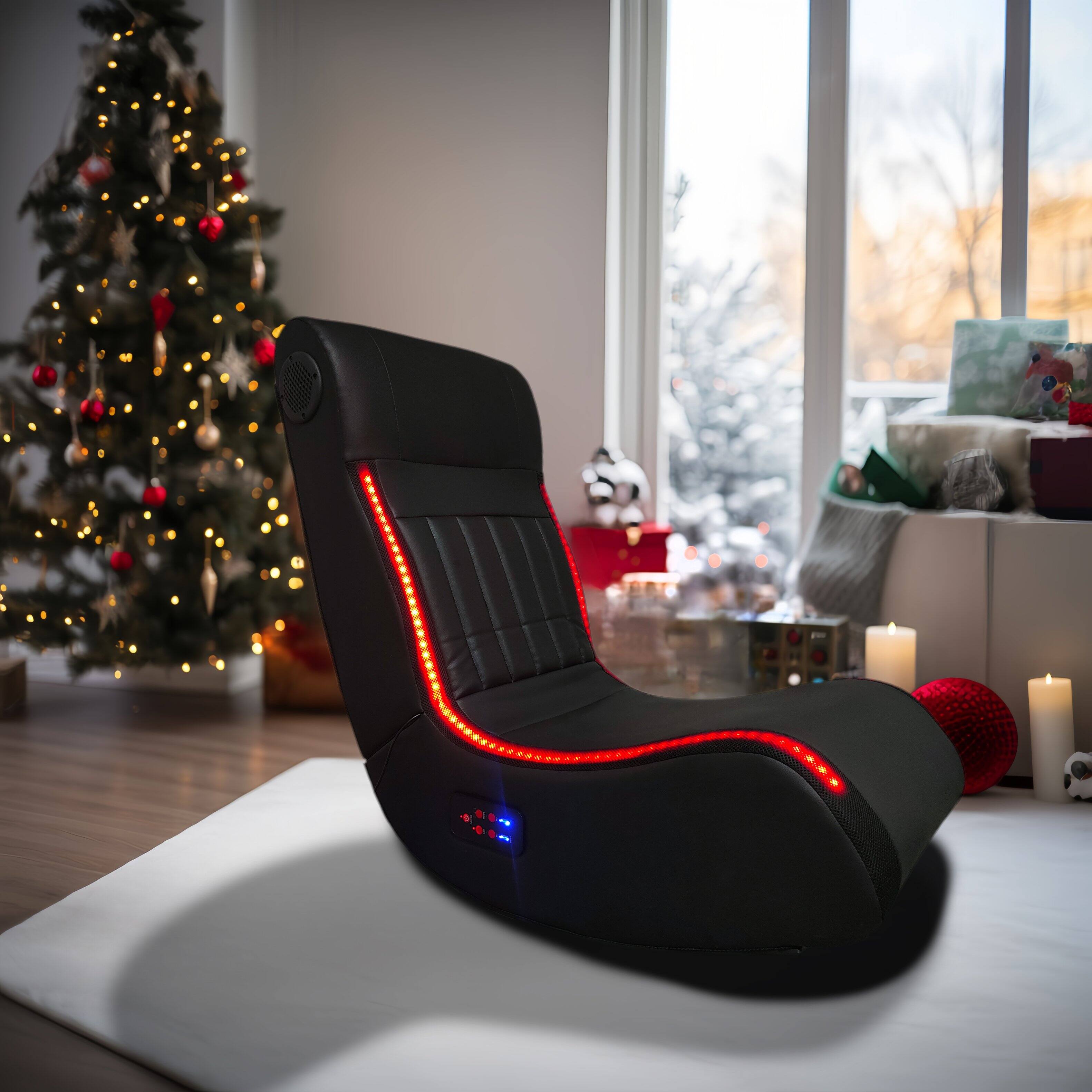 The image shows a modern massage chair with red LED lights along its contours, placed in a cozy room decorated for Christmas. A beautifully lit Christmas tree with ornaments and lights is in the background, along with wrapped gifts and candles, creating a festive atmosphere. The chair is positioned on a white platform, and the room has large windows with a view of a snowy outdoor scene.