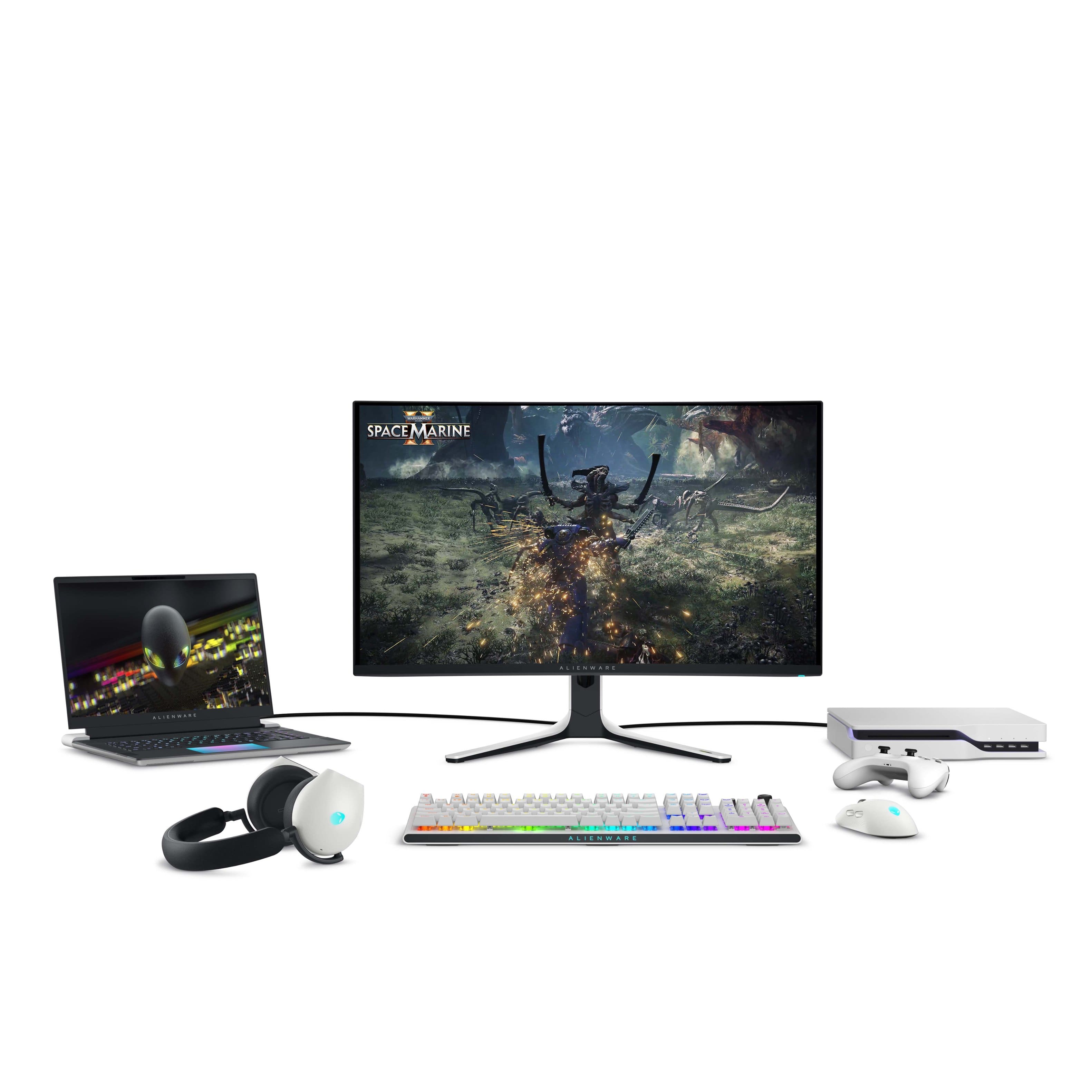 The image features a laptop, a monitor, and a keyboard. The laptop is placed next to the monitor, and the keyboard is positioned in front of the monitor. The monitor is displaying a game, possibly Space Marine. The setup appears to be a workstation or a gaming setup.