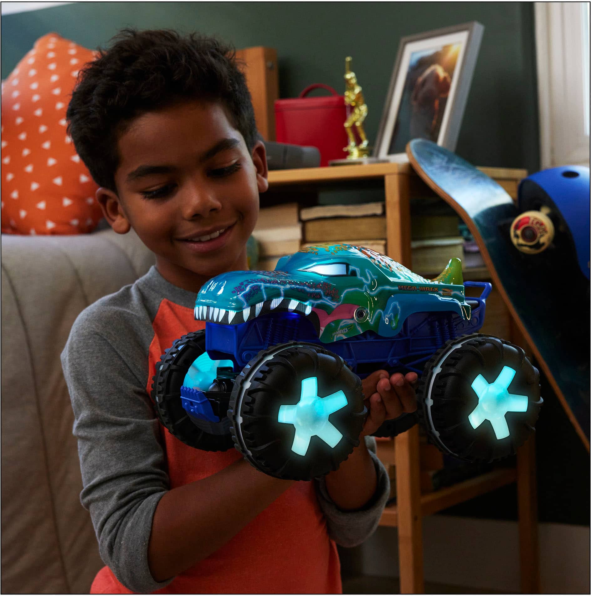 The image shows a young boy holding a blue toy truck with glowing wheels. He is smiling and appears to be proud of his toy. The scene takes place in a living room, with a couch in the background. There are also several books scattered around the room, indicating that the boy may enjoy reading or spending time in this space.