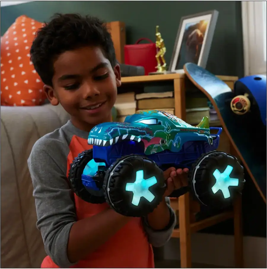 The image shows a young boy holding a blue toy truck with glowing wheels. He is smiling and appears to be proud of his toy. The scene takes place in a living room, with a couch in the background. There are also several books scattered around the room, indicating that the boy may enjoy reading or spending time in this space.