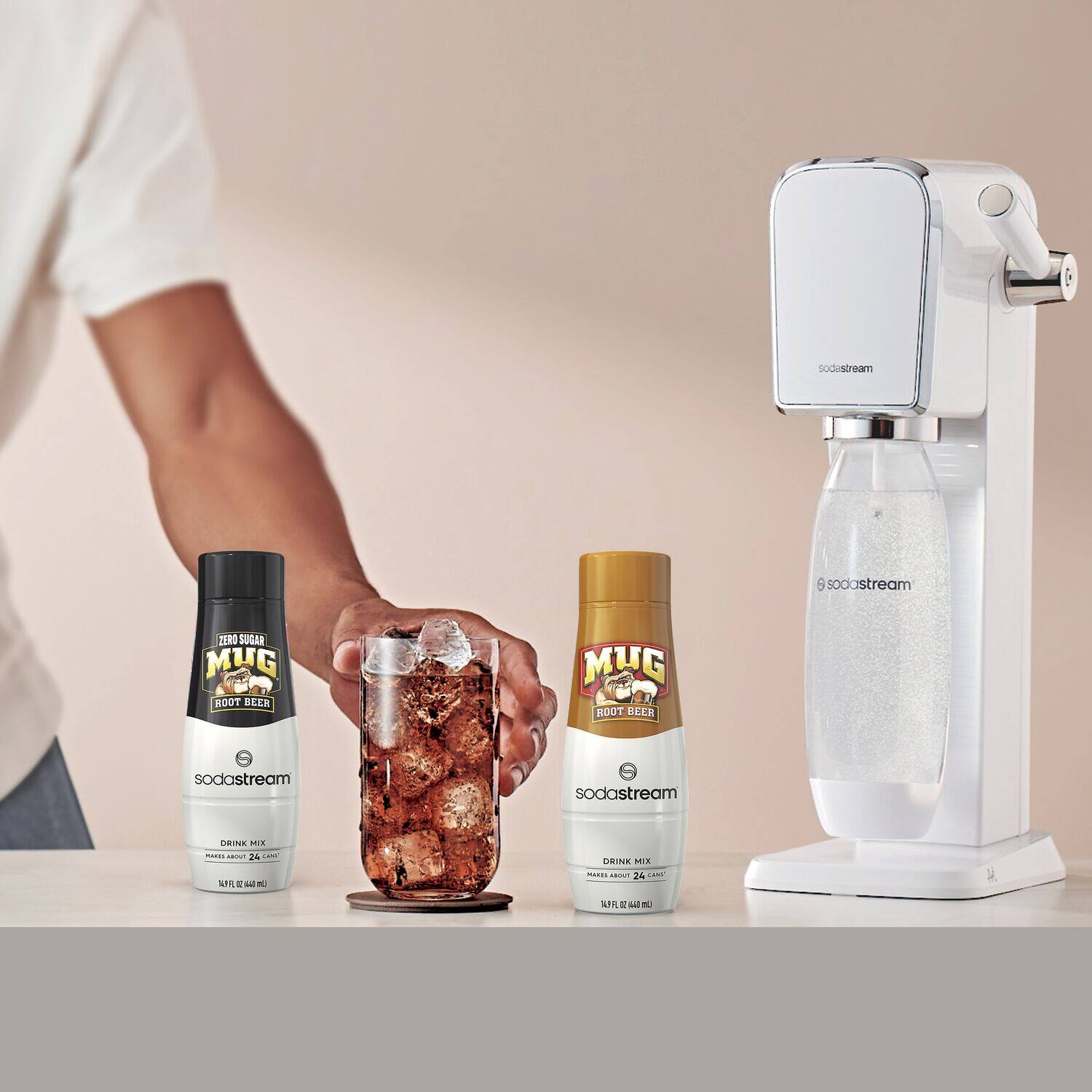 A person is holding a bottle of Mug Root Beer and two other bottles of Mug Soda Stream. The bottles are placed on a table, and there is a Soda Stream machine nearby.