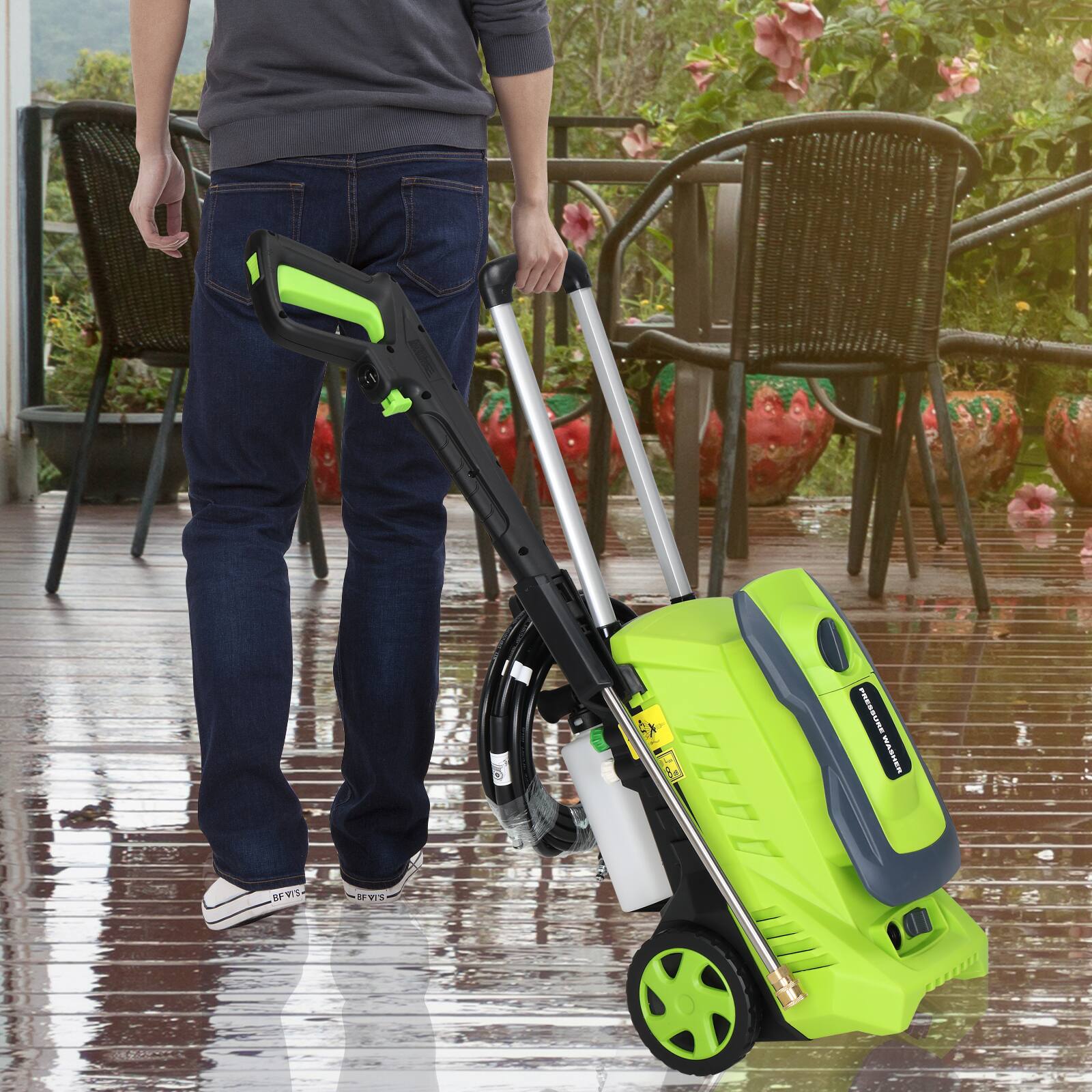 A person is using a green and black pressure washer on a wet wooden deck. The pressure washer has a long handle and a hose attached to it. The person is wearing blue jeans and white sneakers. In the background, there are wicker chairs and potted plants with pink flowers.