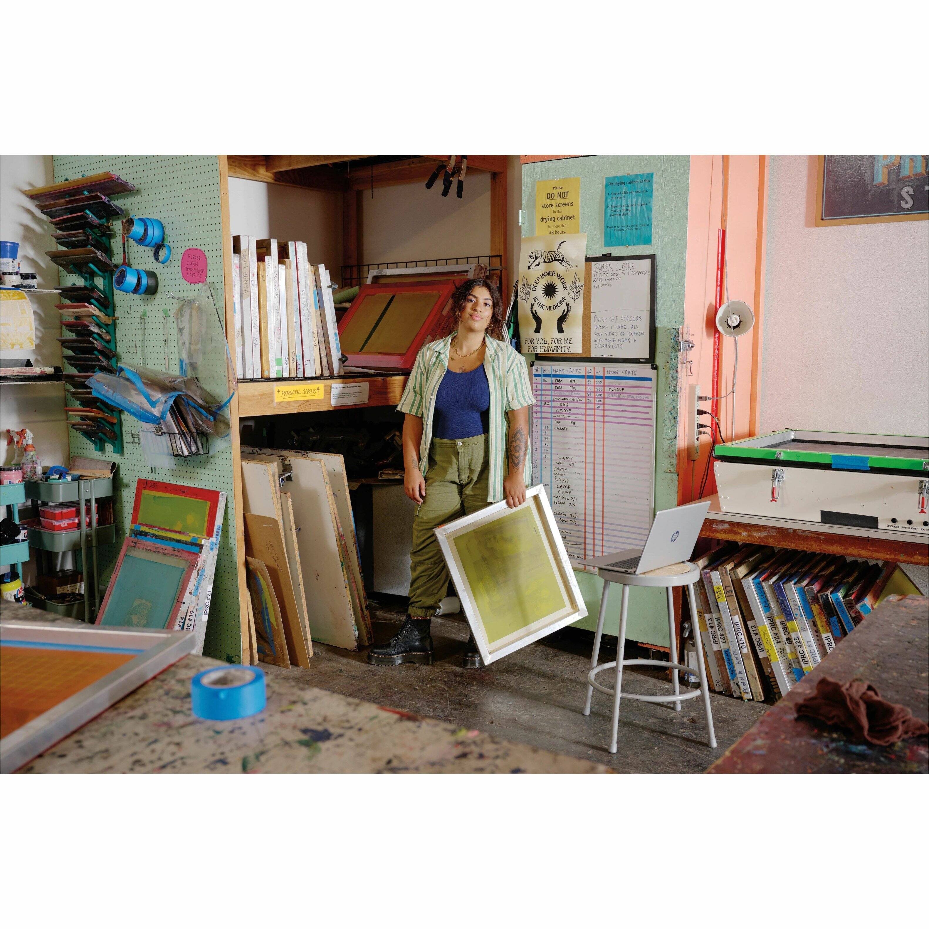 The image shows a woman standing in an art studio, holding a large piece of artwork. She is surrounded by various art supplies, including several books, a laptop, and a chair. The studio appears to be a workspace for creating and displaying art, with a focus on printmaking. The woman is likely an artist or a visitor to the studio, appreciating the artwork and the creative environment.