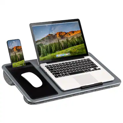 The image shows a laptop computer sitting on a stand, with a cell phone placed on a mouse pad in front of it. The laptop is open and turned on, displaying a beautiful mountain scene on its screen. The cell phone is positioned next to the laptop, suggesting that the user might be multitasking or using both devices simultaneously. The overall scene conveys a sense of productivity and technology integration in everyday life.