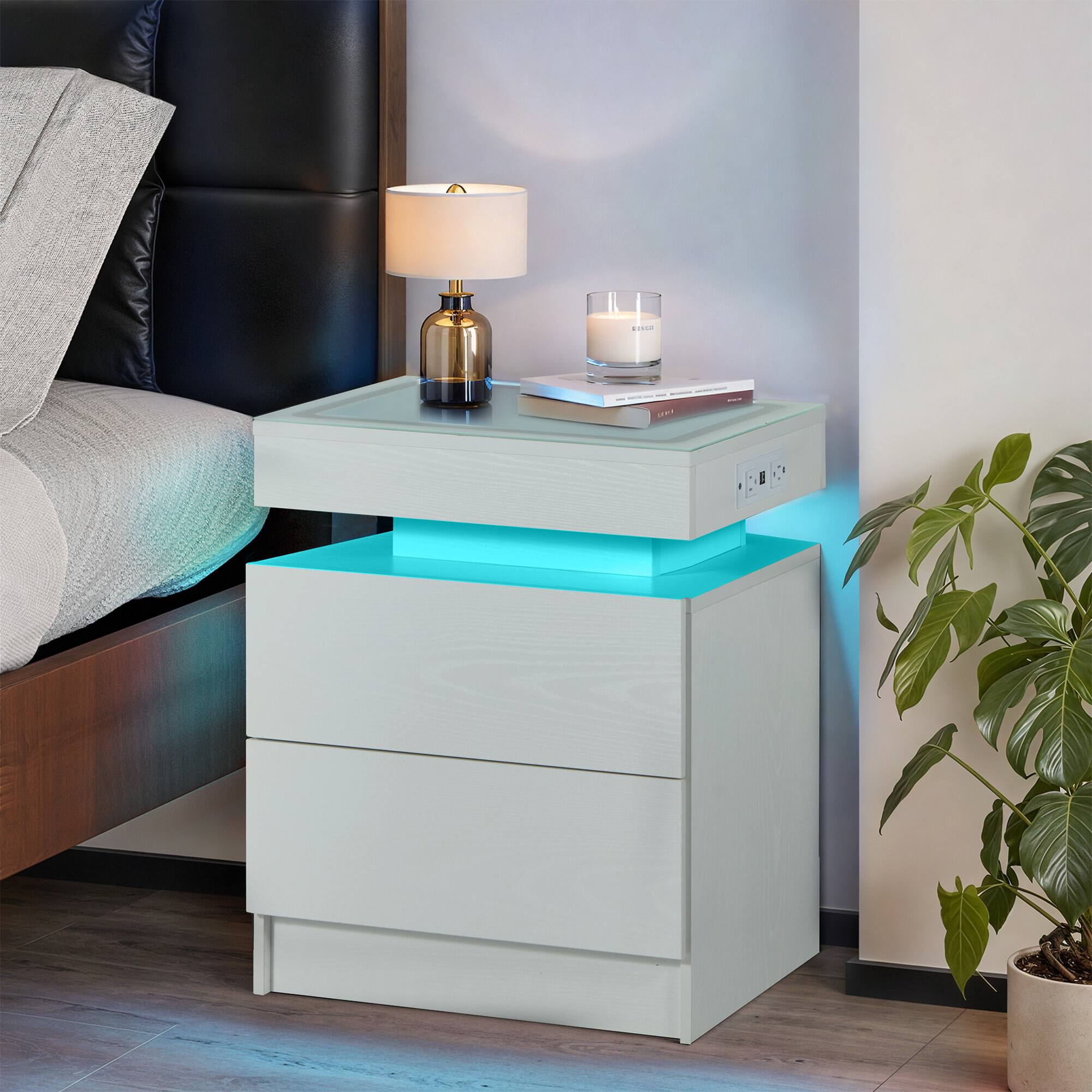 The image does not contain any text. It shows a modern bedside table with a lamp, candle, and books, next to a bed with a plant nearby.