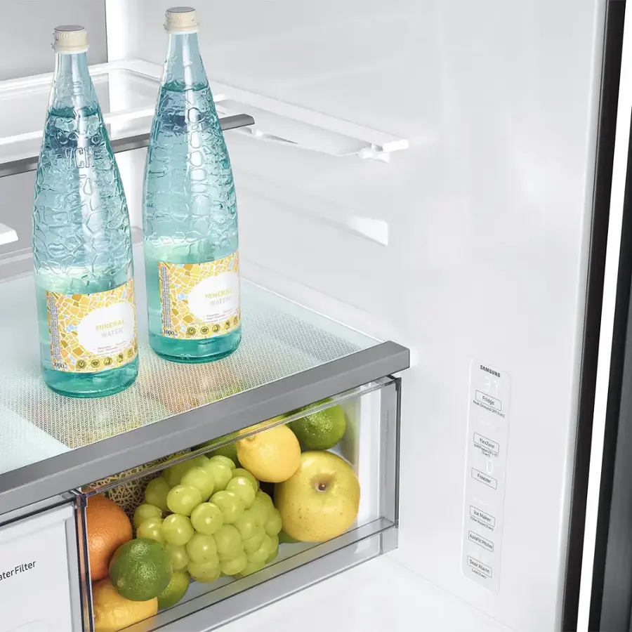 The image shows a refrigerator with two bottles of mineral water placed on the top shelf. The refrigerator also contains a drawer with a variety of fruits, including apples, oranges, and grapes. The apples are located in the middle of the drawer, while the oranges are on the left side and the grapes are on the right side. The refrigerator is a Samsung model, and it has a water filter installed.