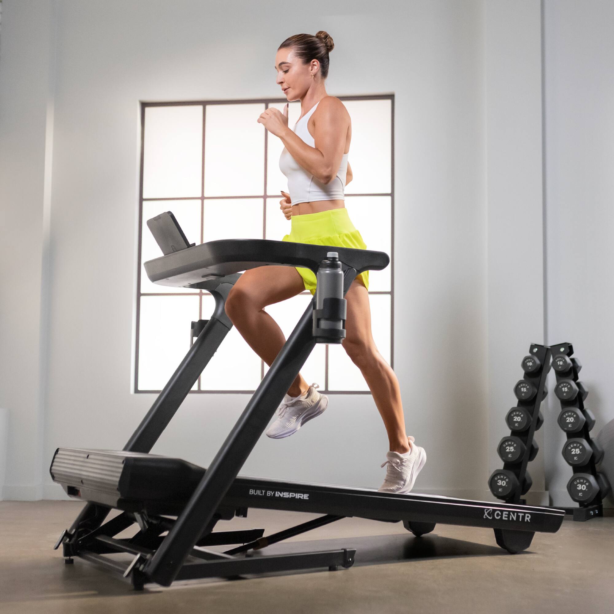1. A woman is running on a treadmill in a gym.
2. She is wearing a white shirt and yellow shorts.
3. The treadmill is black and has a laptop on it.
4. The gym has a window, and there are dumbbells nearby.
5. The woman is running at a speed of 20-25 mph.