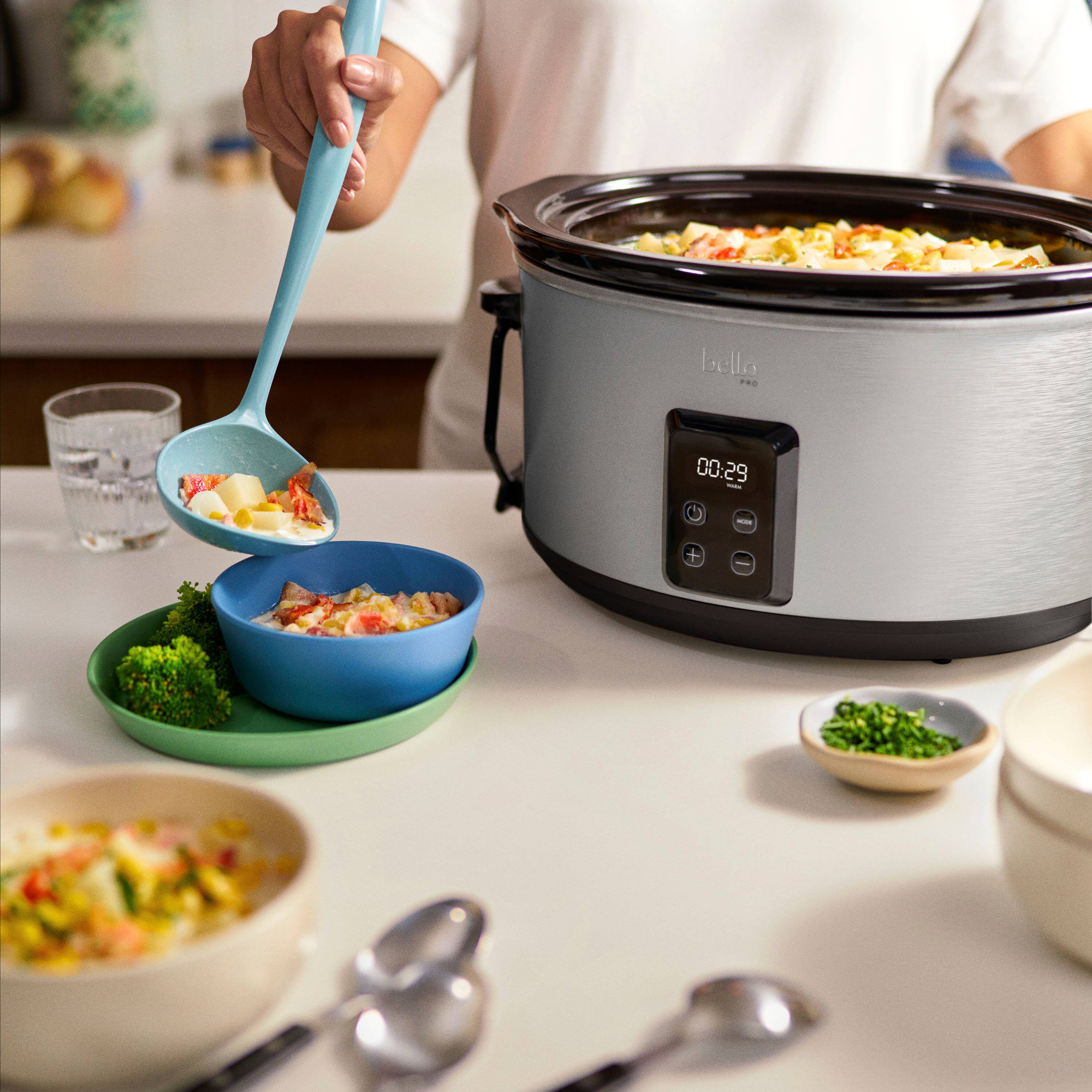 The image shows a person preparing a meal using a Bello crockpot. They are scooping food from a bowl into the crockpot, which is placed on a countertop. The person is also using a spoon to serve the food. The scene includes a dining table with various bowls and cups on it, as well as a spoon and a fork. The person is wearing a white shirt while preparing the meal.