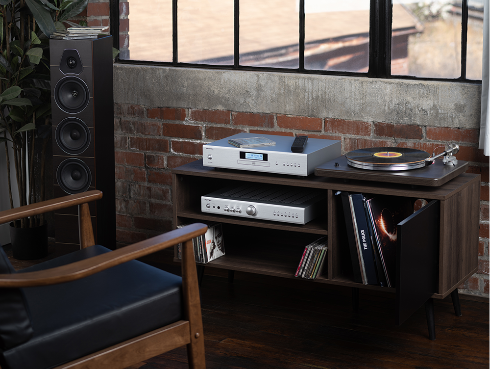 The image shows a room with a black chair, a record player, and a speaker. The record player is placed on a wooden table, and there are several records nearby. The room also features a brick wall and a window, creating a cozy atmosphere.