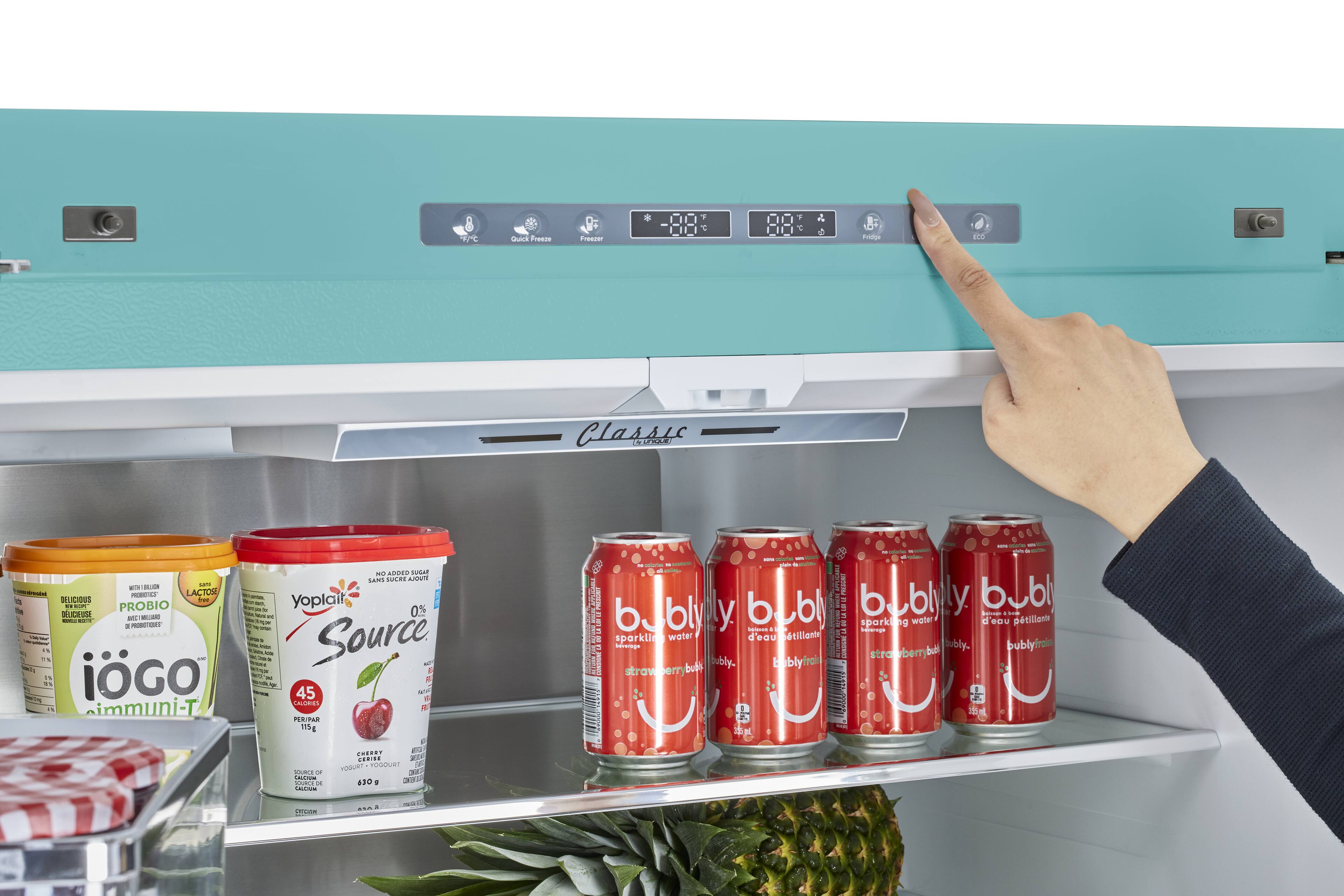 A person is pointing at a refrigerator with a variety of drinks inside, including cans of soda and bottles of water. The refrigerator has a blue door and is stocked with several different beverages. The person is likely selecting a drink to enjoy.