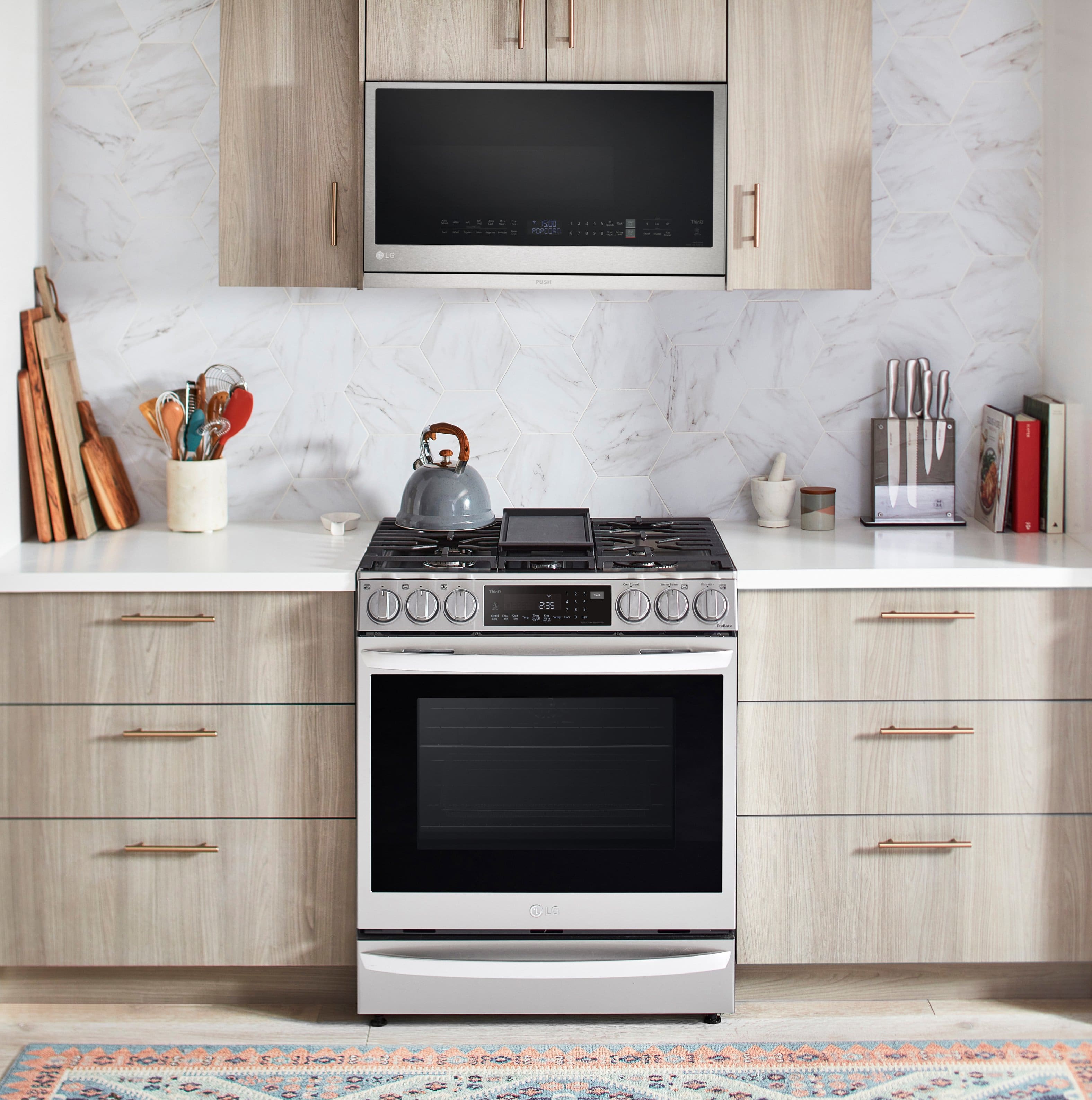 The image shows a kitchen with a stove top oven and a microwave above it. The oven is white and silver, and the microwave is black. The kitchen also features a sink and a countertop. There are several knives and a spoon placed on the countertop, along with a few books. The scene is set in a kitchen with a tile backsplash and a rug on the floor.