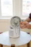 The image shows a person holding a small appliance, possibly a fan or a heater, while standing near a window. The appliance is white and appears to be a portable device. The person is likely trying to operate or control the device, possibly to adjust the temperature or airflow. The scene takes place in a room with a dining table and a chair, suggesting that it might be a living or dining area.