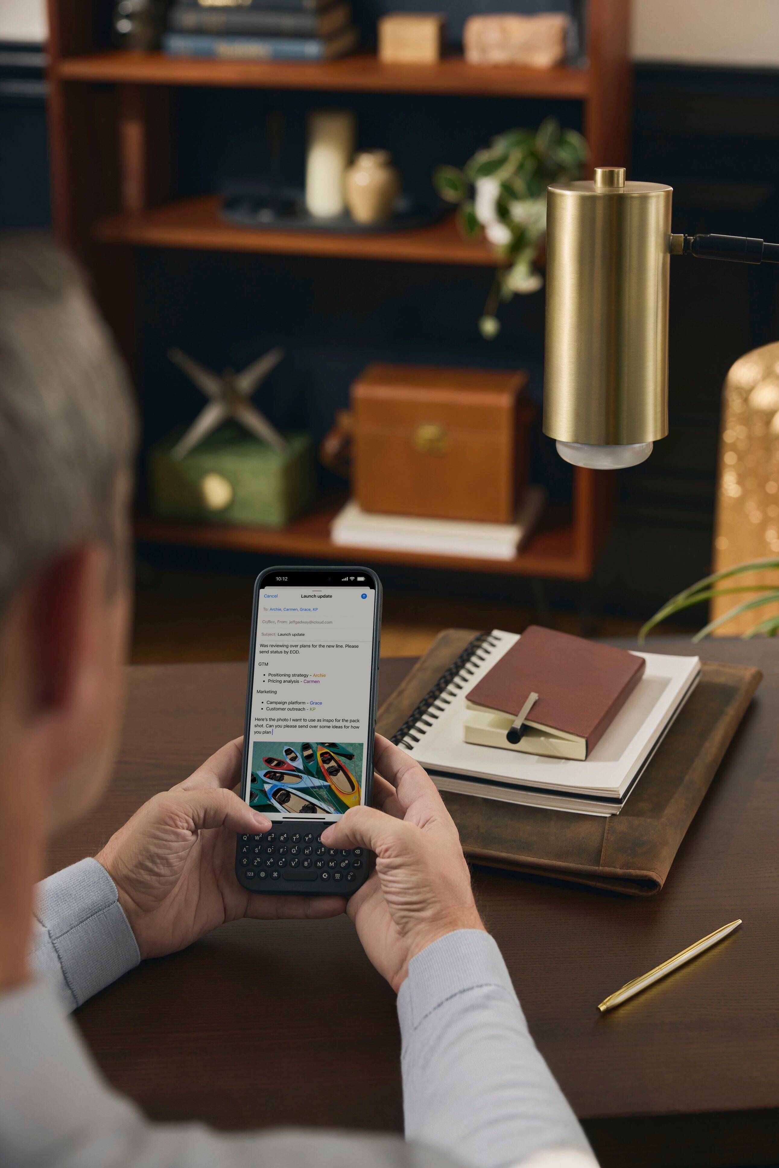 The man is sitting at a desk and looking at his cell phone. He is holding the phone in his hands, possibly texting or browsing the internet. The desk is cluttered with various items, including a book, a vase, and a potted plant. There is also a remote control on the desk, and a clock is visible in the background. The man appears to be focused on his phone, possibly checking his messages or working on a task.