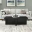 The image features a living room with a black coffee table in the center. The table has a book on top of it, and there are two pictures hanging above the couch. The couch is white, and there are two pillows on it. The room is decorated with a rug, and there are two lamps on either side of the room. The overall atmosphere of the living room is cozy and inviting.